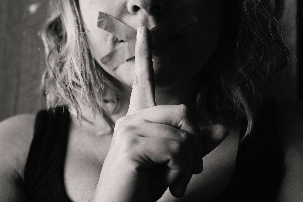 Scared woman with gag over mouth and finger up to her lips, beckoning you to be quiet.