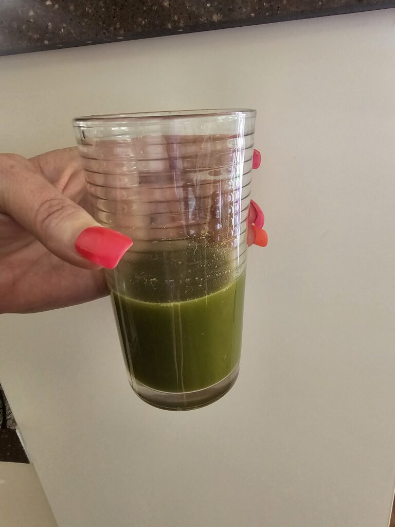 Womens hand with long pink fingernails holding glass cup containing Barley Grass