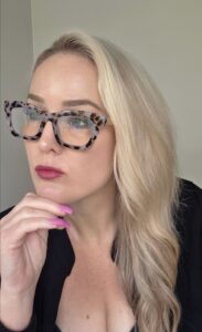 Blonde haired woman wearing reading glasses sits thoughtful, contemplating life