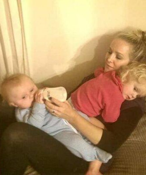 Blonde woman sitting on couch with toddler cuddling her and baby sitting on her lap, drinking her bottle.